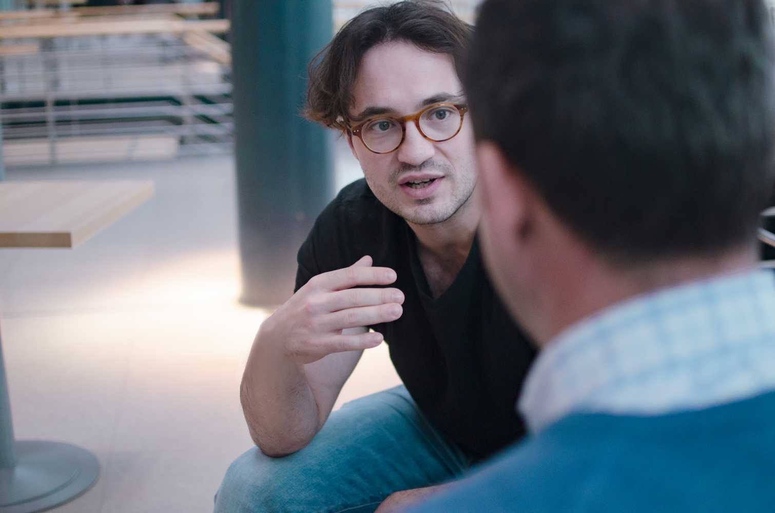 Man with glasses in the background is talking to a person in the foreground
