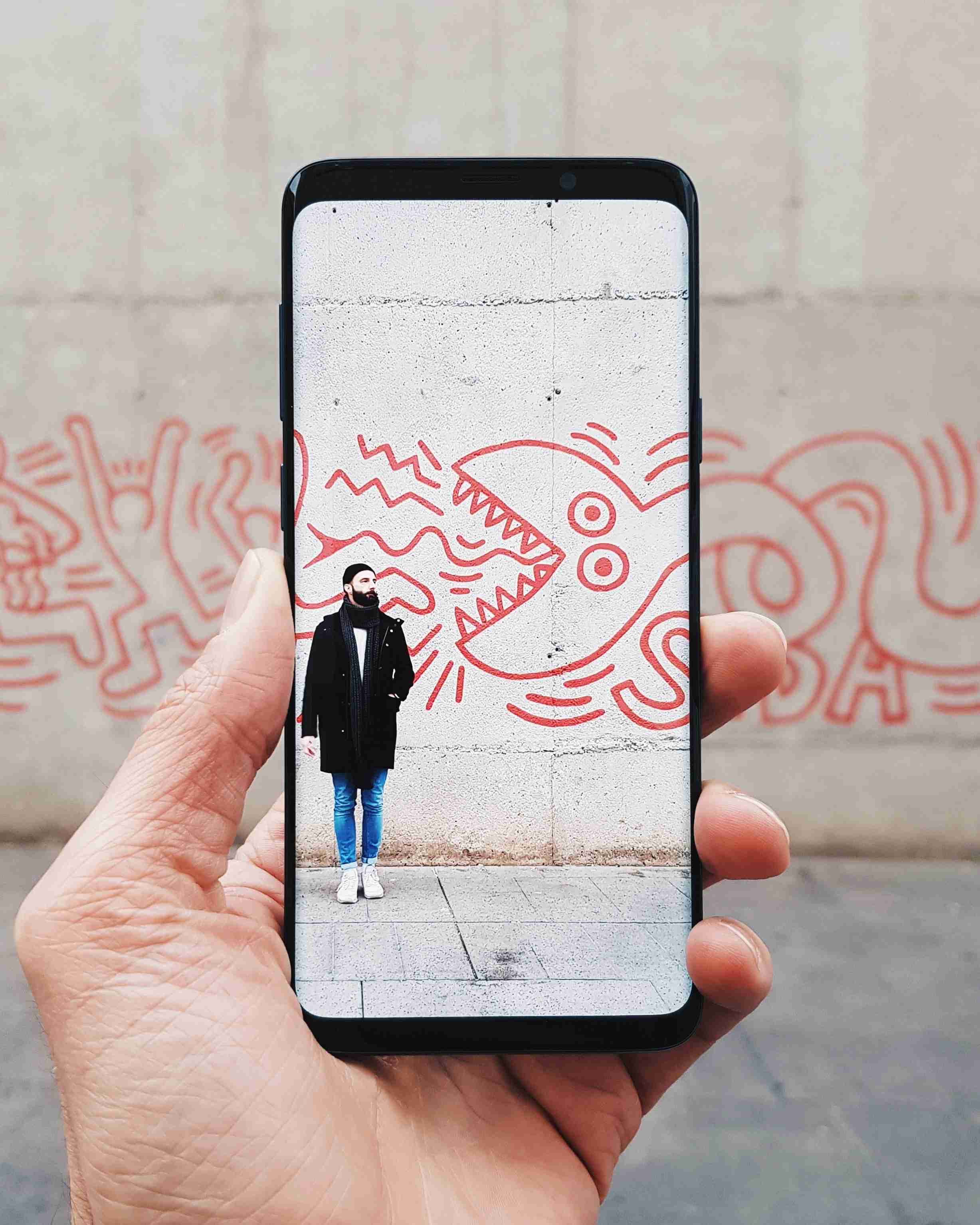 Foto von einem Handy auf dessen Bildschirm ein Mann bei einem Graffiti stehend zu sehen ist.